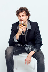 Confident young man in a jacket sitting on the chair and holding glass of wine over white background