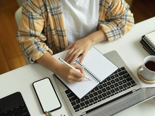 Female in Scott jacket hand writing on blank notebook and working with smartphone and laptop on the table