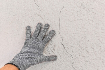 Hand of a working man in a construction glove examines cracks on a white wall background. Repair concept