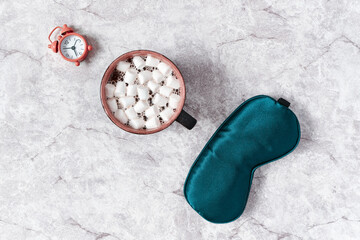 Silk sleep mask, alarm clock and a cup of cocoa with marshmallows are on a gray background. The concept of good sleep, waking up in the morning, breakfast and planning the day. Flat lay.