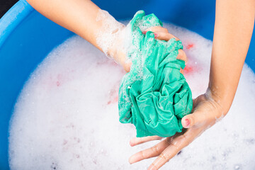 Fototapeta premium Closeup young Asian woman use hands washing color clothes in basin. Female squeeze wring out wet fabric cloth with detergent have soapy bubble in water, studio shot background, laundry concept