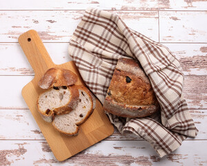 Rustic whole grain artisan bread loaf slice with cranberry raisin dry fruit nuts wrapped in checkers cloth with wooden chopping board over table top flat lay view