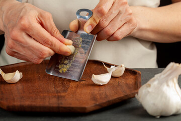 Hands of a caucasian woman grating garlic cloves using shovel shaped metal mini grater on a wooden plate. This small item can also be used as ginger grater. A convenient durable tool.