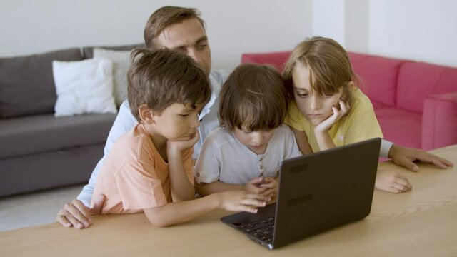 Serious dad and kids watching movie on laptop together. Middle-aged father sitting at table with children. Boys and girl looking at screen. Hand-held camera. Fatherhood and digital technology concept