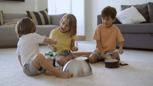 Happy little kids playing in living room with utensils. Blonde girl and two boys smiling, sitting on floor together and knocking on pans. Static camera. Childhood, noise and home activity concept