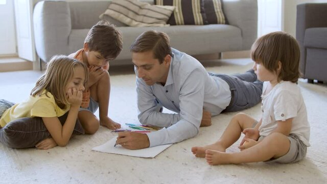 Sibling kids and dad sitting and lying on warm floor with cushions, playing together, drawing and talking. Childhood or parenthood concept