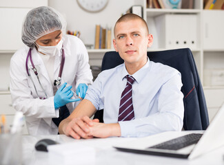 Doctor in white coat and protective mask giving vaccine injection to manager in office