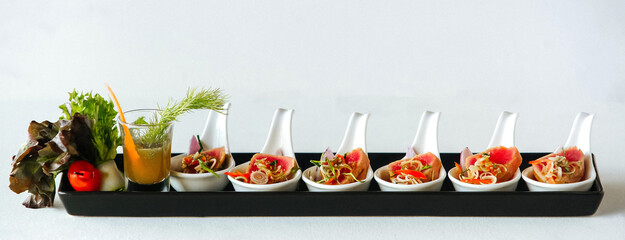 Snacks in white spoons with red potatoes or meat, with vegetables and sauce in a shot glass on long plate on white background 