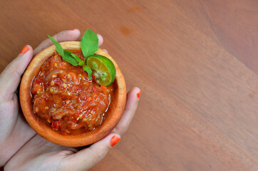 Asian female hands give chilli paste (sambal terasi) in a wooden bowl with garnished basil leaves and lime wedges. selective focus