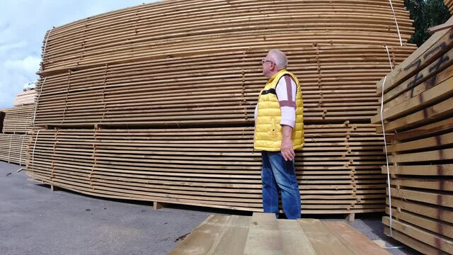 Senior man in glasses and yellow tank top and jeans examines industrial wooden boards in open warehouse under blue sky