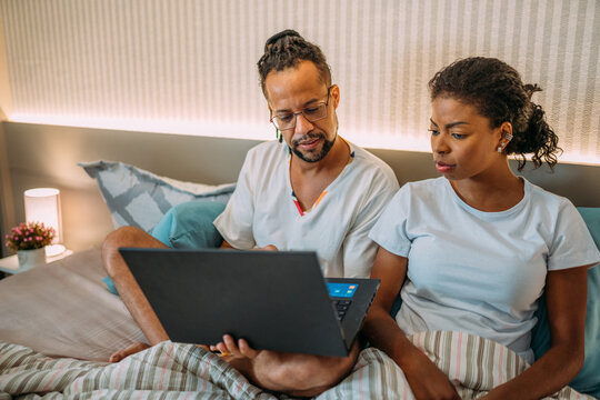 Latinx Couple Lying In Bed, Husband Showing Something To Wife On Laptop