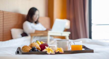 Obraz na płótnie Canvas Focus on sweets fruit. Young Asian women smiling happily at freelance work, working on a notebook while relaxing in bed with snacks and fruit in a hotel room. Vacation and relaxation