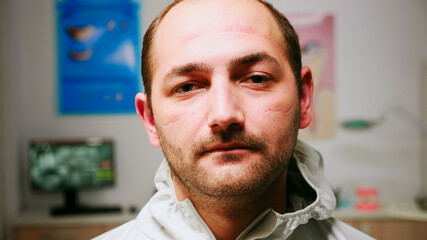 Obraz premium Close up of tired pediatric stomatologist man taking off face shield looking at camera exhausted sitting in new normal dental office. Medical team wearing ppe suit and gloves