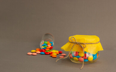 Chocolate candies, small multi-colored in a jar with a bow, on a gray background and with a yellow lid.