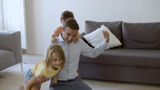 Exited Caucasian man playing with children and showing strength. Cheerful kids having fun together in living room on carpet. Hand-held camera. Front view. Childhood, weekend and home activity concept