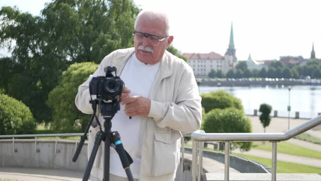 Senior man in white jacket adjusts camera on tripod on embankment against green trees river buildings and church tower closeup