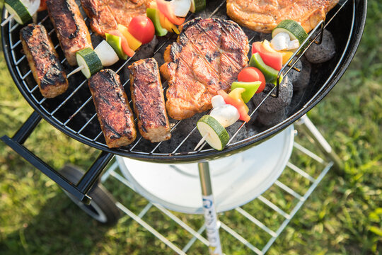 Marinierte Schweine Fleisch Nackensteaks und bunte Gem&uuml;sespie&szlig;e grillen mit Rauch auf Holzkohle Kugelgrill im Garten an Sommer Abend