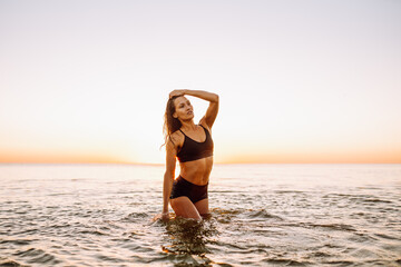 Fototapeta na wymiar Young sporty woman with beautiful slim body and long dark hair posing in calm sea at sunset in black sportswear. Space for message