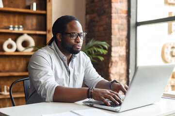 Fototapeta na wymiar Busy bored young African-American male author office worker in glasses sitting at the desk typing on laptop, working on project, writing creating article or blog, feeling lack of motivation to finish