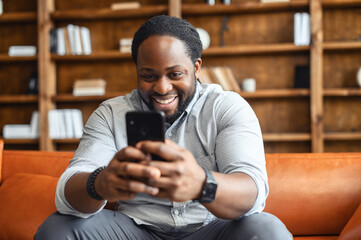 Happy laughing mixed-race man sitting on the couch, staying at home, texting chatting with friends...