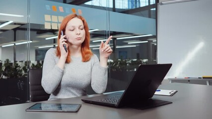 Red haired woman talking on phone in business office, using laptop, tablet lying beside, Agile board on glass wall behind her. Consultation of financial advisor. Concept of communication