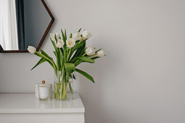 White tulips in a glass vase on a white chest of drawers.