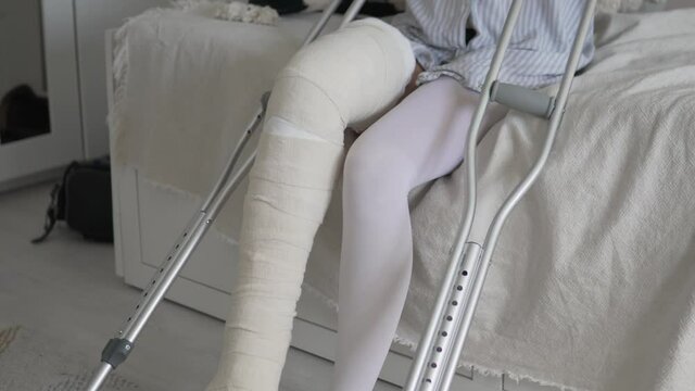 Close-up of the legs in high plaster, a woman on crutches to sit on the bed.