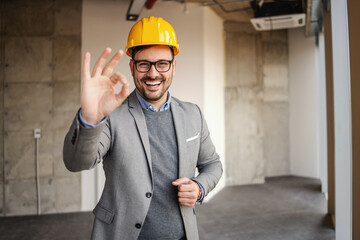 Smiling architect standing in building in construction process and showing okay gesture.