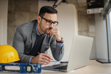 Focused architect leaning on desk at construction site with pencil in his hand and looking at...