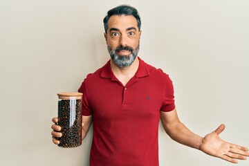 Middle age man with beard and grey hair holding jar with coffee beans celebrating achievement with happy smile and winner expression with raised hand