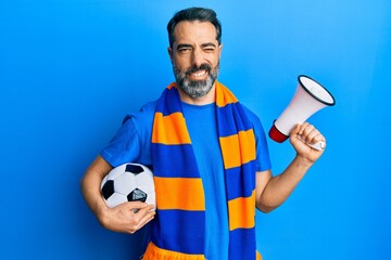 Middle age man with beard and grey hair football hooligan holding ball and using megaphone winking looking at the camera with sexy expression, cheerful and happy face.