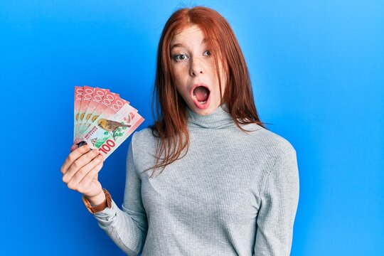 Young red head girl holding 100 new zealand dollars banknote scared and amazed with open mouth for surprise, disbelief face