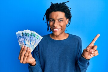 Young african american man holding south african 100 rand banknotes smiling happy pointing with...