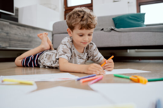 A school-age boy lies on the living room floor and paints pictures on white sheets of paper with colored markers. The boy is spending his afternoon free time at home.