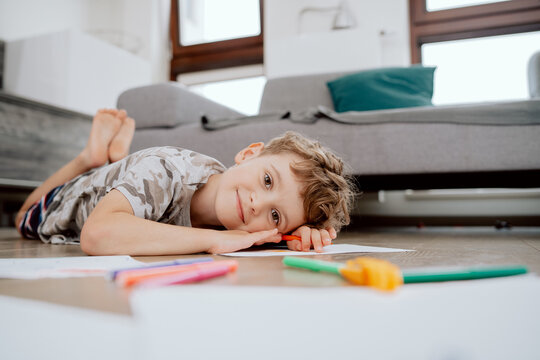 Portrait of school-age boy lies on the living room floor and paints pictures on white sheets of paper with colored markers. The boy is spending his afternoon free time at home.