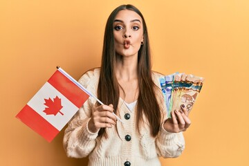 Beautiful brunette young woman holding canada flag and dollars making fish face with mouth and...