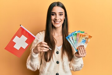 Beautiful brunette young woman holding switzerland flag and franc banknotes smiling with a happy and cool smile on face. showing teeth.