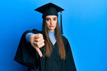 Beautiful brunette young woman wearing graduation cap and ceremony robe looking unhappy and angry...