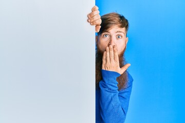 Young irish redhead man holding blank empty banner covering mouth with hand, shocked and afraid for...