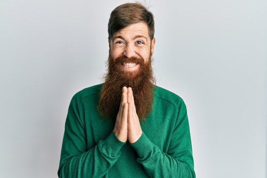 Redhead man with long beard wearing casual clothes praying with hands together asking for forgiveness smiling confident.