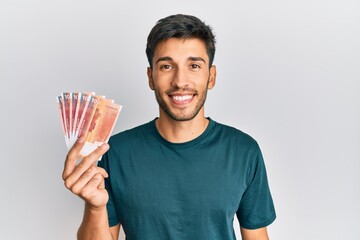 Young handsome man holding 100 norwegian krone banknotes looking positive and happy standing and smiling with a confident smile showing teeth