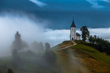Jamnik, Slovenia - Foggy summer morning at Jamnik St.Primoz hilltop church. The fog gently goes around the small hilltop chapel with green fields and animals on the top of the hill - obrazy, fototapety, plakaty