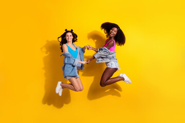Fototapeta na wymiar Photo of two adorable pretty young ladies wear jeans outfit holding arms jumping arms isolated yellow color background