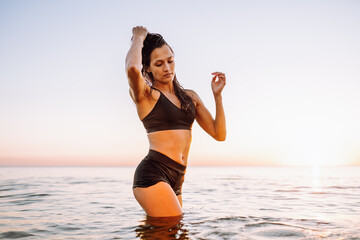 Young sporty woman with beautiful slim body and long dark hair posing in calm sea at sunset in black sportswear. Space for message