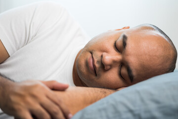 An Asian bald man around the age of 30 in a white T-shirt is sleeping on his side on a blue mattress and pillows. He slept at home until morning.