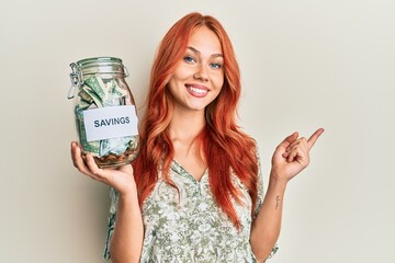 Young beautiful redhead woman holding charity jar with money smiling happy pointing with hand and finger to the side