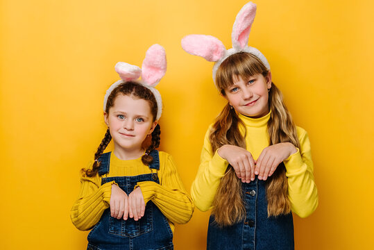 Two charming little girls keeps hands as if rabbit, wears soft bunny ears, has friendly look, ready to congratulate children with coming Easter, models on yellow studio background. I am lovely hare