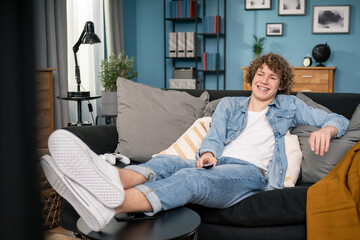 Obraz premium Teen boy sitting on cozy couch and watching comedy on TV laughing with humorous jokes.