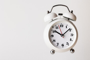 Small white alarm clock, black numbers, set the time for 10.10 o'clock, placed on a white table.