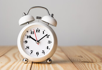 Small white alarm clock, black numbers, set the time for 10.10 o'clock, placed on a wood table.